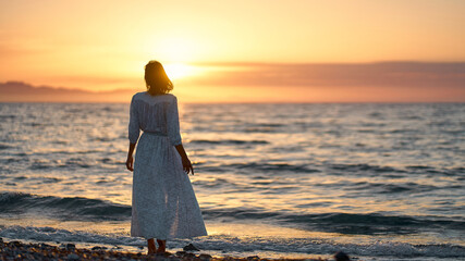 Happy carefree woman in long dress enjoying beautiful sunset on the beach. Silhouette of alone girl looking at the horizon