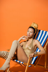 Trendy african american model in swimsuit and knee boots posing on deck chair on orange background.