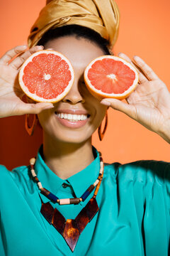 positive and stylish african american model covering face with grapefruit on orange background.