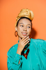 Smiling african american woman in golden headscarf touching chin on orange background.