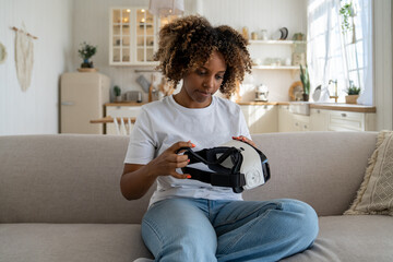 Millennial African American girl with curly hair sitting on couch testing new VR headset, using virtual reality helmet to view photos and videos in 3D. Young woman examining AR glasses at home