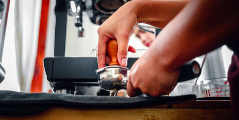 Hand of a barista holding a portafilter and a coffee tamper making an espresso coffee. Barista presses ground coffee using a tamper in a coffee shop