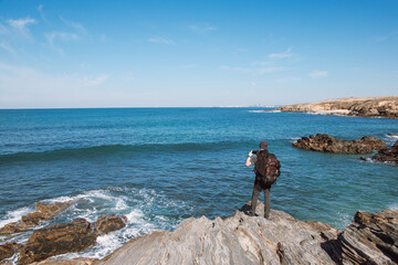 Elderly man with a backpack photographs the beauty of the Atlantic coast near Porto Covo, Portugal. Capturing moments on a mobile phone