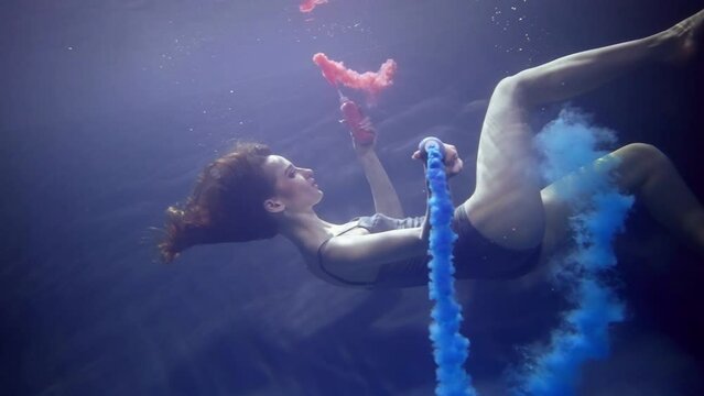 beautiful young woman diving in depth of pool slowly, painting water by red and blue dyes in tubes