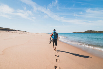 Passionate backpacker, backpack and boots in hand, walks along the Praia da Ilha do Pessegueiro beach on the Atlantic Ocean near Porto Covo, Portugal. In the footsteps of Rota Vicentina