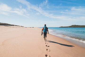 Passionate backpacker, backpack and boots in hand, walks along the Praia da Ilha do Pessegueiro beach on the Atlantic Ocean near Porto Covo, Portugal. In the footsteps of Rota Vicentina © Fauren