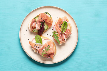 Set of bruschetta with jamon or prosciutto, vegetables and fresh arugula leaves on the plate on bright blue background top view. Healthy homemade breakfast or lunch idea.
