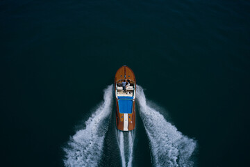 Big luxury wooden motor boat with man and woman moving faster on dark water top view from behind. Large motor boat with people moving on the water, making a white trail, aerial view. - obrazy, fototapety, plakaty