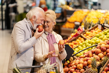 An old man is biting fresh apple at the supermarket while his wife is looking at him.