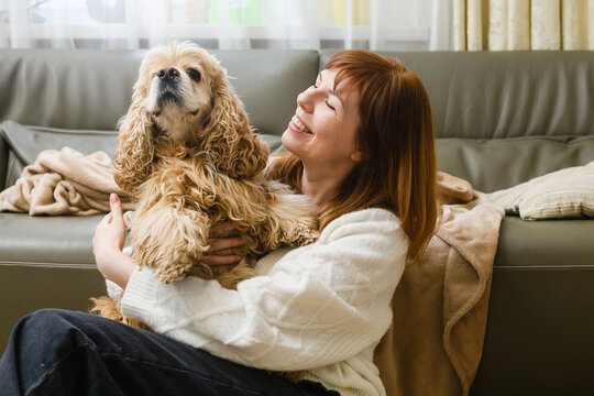A young woman is holding a Cocker Spaniel in her arms while sitting on the floor near the sofa.