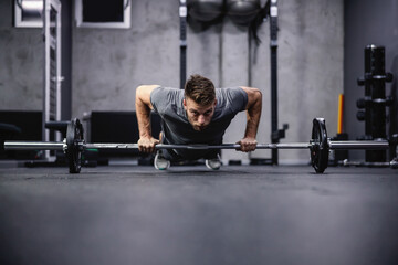 Obraz na płótnie Canvas Push-ups and muscle strengthening. An attractive and muscular man does push-ups leaning his hands on a barbell. A man in sportswear pumps his arm muscles in a modern gym. Sport, fitness challenge