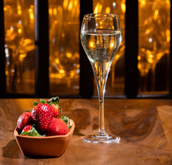 Glass of brut champagne bubbles wine in tulip glass and bowl with fresh strawberries on evening bar lights background