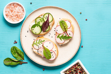 Bright juicy spring and summer snack. Set of bruschetta with cream cheese, cucumbers, small radish and fresh salad leaves on light blue background. Healthy homemade food ideas.