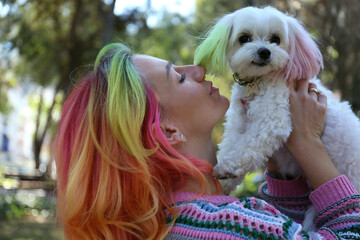 Close up shot of a young woman with multicolored hair kissing her beloved maltese dog. Female and her pup with color matched ears. Copy space, background.