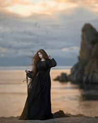 Girl in a black dress with a train with a sword on the seashore at sunset