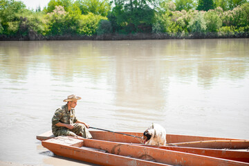 Bored fisherman in hat going fishing, while sitting in rowing old boat at shore in morning. Side view of caring man in camo looking at cute dog, while angling in calm river. Concept of rest, pet care.