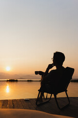 Fototapeta na wymiar Silhouette tourist man sit on a chair holding cup of coffee wtaching sunrise.