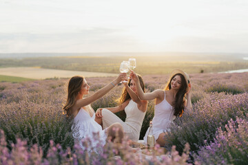 Three happy women toasting with wine while sitting at picnic on the lavender field at sunset. 