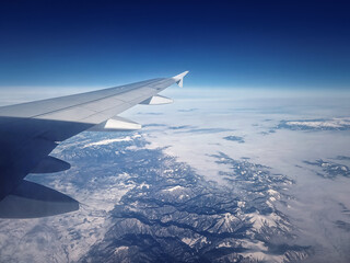 Fototapeta na wymiar Plane flight above the Carpathian Mountains snowy peaks. Blue skyline and airplane wing seen through the window