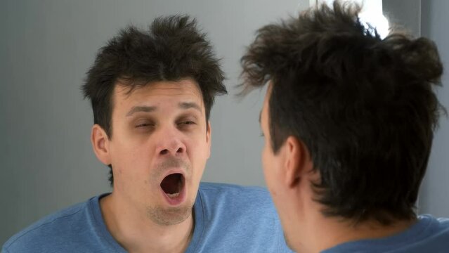 Tired man with a hangover looks in the mirror at his reflection yawns in the morning. He rubs his head and hair and looks at his reflection in the mirror with light bulbs. Consequences of drinking.