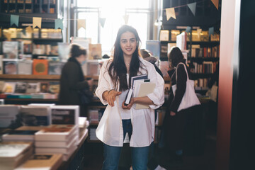 Smiling woman with books looking at camera in bookstore - obrazy, fototapety, plakaty
