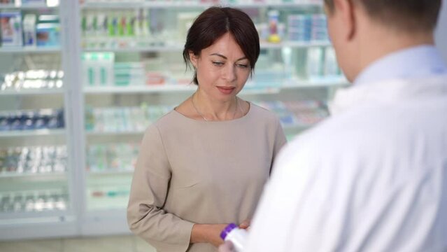 Portrait of Caucasian woman listening to pharmacist advising pills smiling in slow motion. Beautiful female customer in drugstore with professional man passing bottle of medication