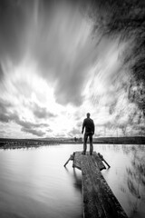 Vertical grayscale shot of a person on a boardwalk by the lake