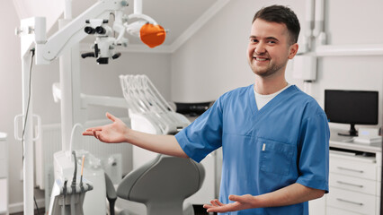 Naklejka na ściany i meble Handsome young dentist in white coat is looking at camera and showing his office