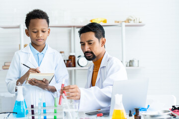 In chemistry classroom with many laboratory tools on table. A young African boy and male teacher in white lab coat doing experiment together. A teacher checking at red test tube and boy writing data.
