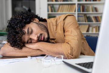 Tired student sleeping on table inside university academic library, hispanic man among books with...