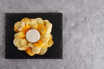 Top view of heap of potato chips with creamy white dipping sauce on black slate serving board on...