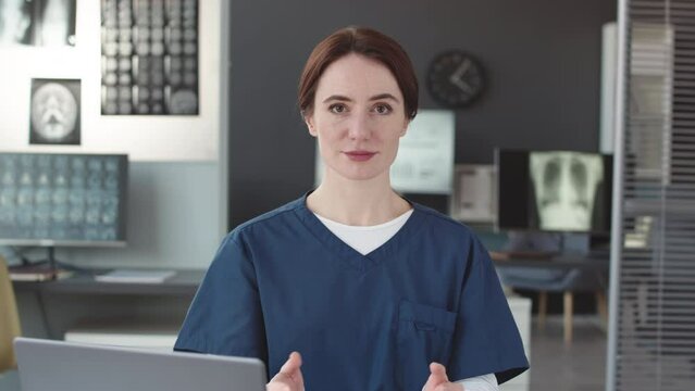 Waist up POV of young Caucasian female doctor in blue scrubs looking and gesticulating at camera sitting at desk in modern office