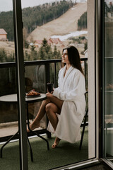 A woman in a white bathrobe eats a croissant and drinks coffee in a hotel in the mountains.Resort vacation concept