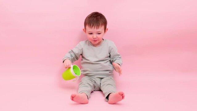 Happy toddler baby drinks water from a non-watering glass on a studio pink background. A smiling child with a green no spill plastic cup in his hands. Kid age one year eight months, full height