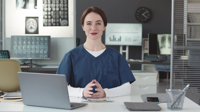 Waist up POV of young Caucasian female doctor wearing scrubs sitting at desk in contemporary doctor office looking and speaking at camera