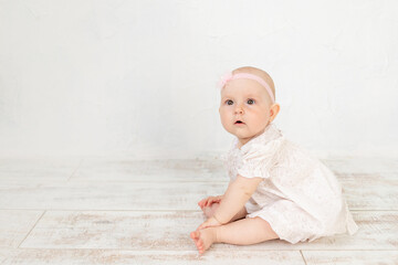 cute baby girl in a white dress sits on the floor for six months in a bright room, a place and space for text