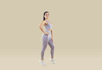 Full-length portrait of smiling active woman athlete in sportswear posing on nude brown studio background. Toned fit girl coach or trainer show good body shape. Fitness and sport concept.