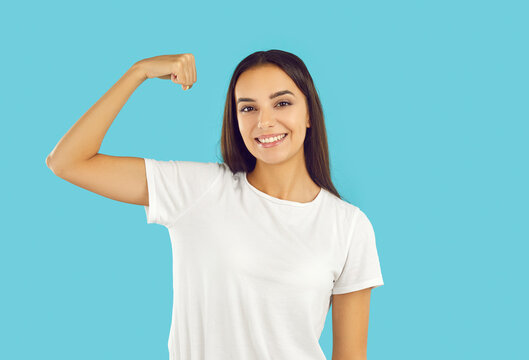 Happy beautiful young woman showing she is strong, healthy and confident. Female model in white mockup T-shirt standing isolated on blue background, flexing arm and smiling. Girl power grl pwr concept - Powered by Adobe