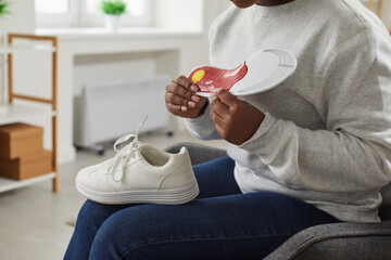 Cropped shot of a young African American woman with a sneaker on her lap sitting in an armchair at home and holding an orthotic arch support shoe insole in her hands. Footwear, feet health concept - obrazy, fototapety, plakaty