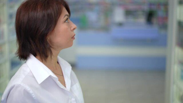 Concentrated woman choosing vitamin supplements on shelves of pharmacy as live camera panning around thoughtful focused female client. Caucasian buyer examining medications in drugstore indoors