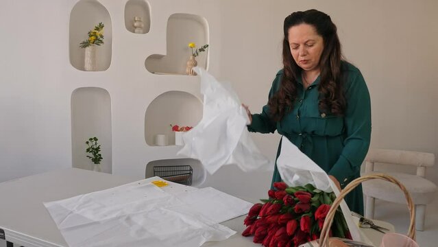 Caucasian lady florist collects a lush bouquet of tulips. A professional makes a fresh bouquet. Small business. A woman crumples paper (Crepe) and wraps a bouquet of tulips in it