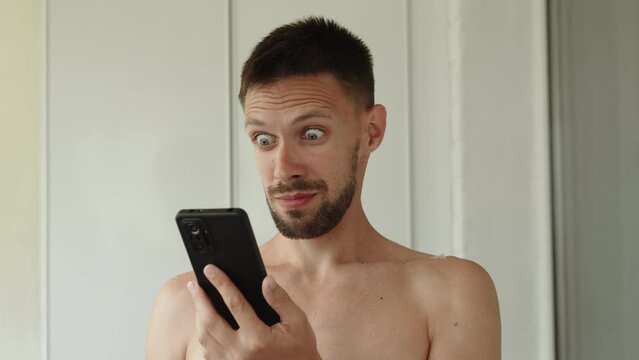 Naked thin man stands in room and looks at phone screen. Man eyes widen slowly, guy is very surprised. Person with beard in stress, excitement and shock from news, closes his eyes with hand.