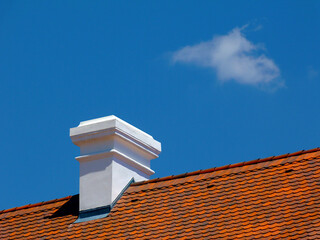 white stucco finished chimney. sloped roof with red brown clay roof tiles. metal roof flashing. blue sky and white cloud. bright sunlight. building construction and renovation concept.