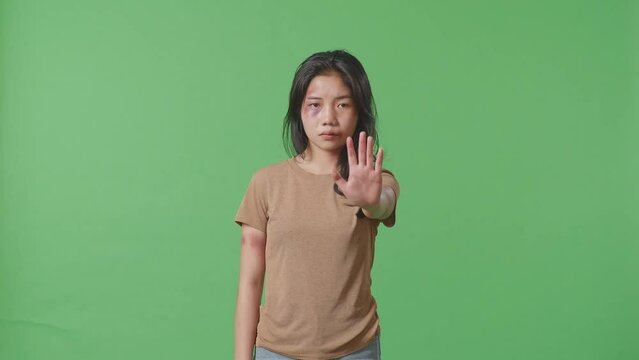 Young Asian Woman With Bruise On Face And Arms Looking Into Camera Showing Hand Sign To Stop Violence On Green Screen Background In The Studio
