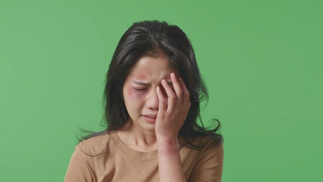 Close Up Of Young Asian Woman Victim Of Violence With Bruise On Face Covering Her Hurt Eye And Crying On Green Screen Background In The Studio
