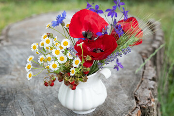 Blurred image of a field bouquet with poppies, daisies, wild strawberries and spikelets on a stump.