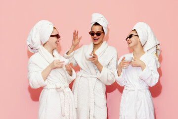 Three cheerful pretty young girls in bathrobes and towels, drinking coffee after spa against pink studio background. Concept of youth, face care, beauty, friendship, party, relaxation