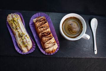 Cup of coffee and delicious eclair cakes - tiramisu and pistachio flavoured covered with cream, icing and chopped nuts, on a black background.