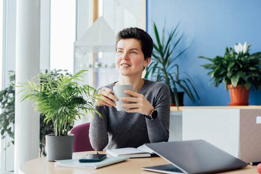 Satisfied smiling newcomer middle aged neutral gender female employee holds coffee cup and sitting at her new office desk work space. First working day concept. Dreaming about new opportunities. - Powered by Adobe