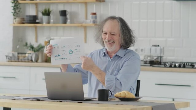 mature man is having a video conference at home in the kitchen showing financial reports on paper. Online remote work concept